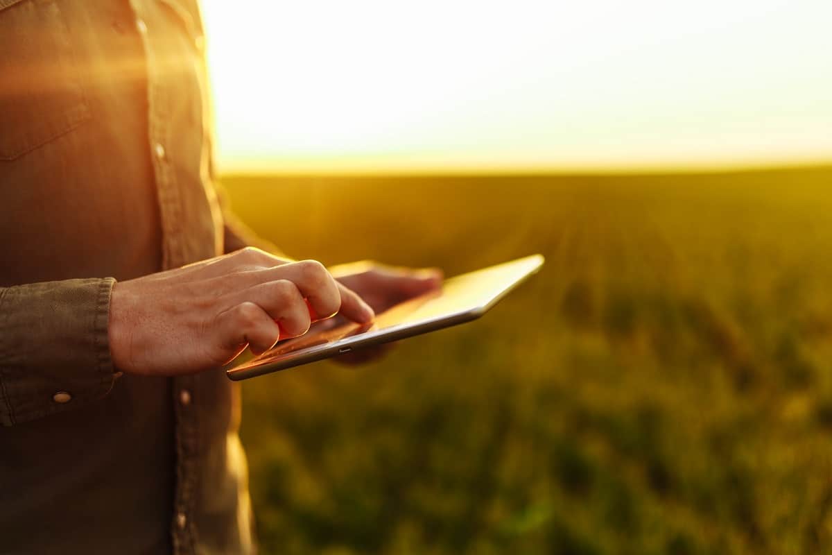 Foto de homem segurando um tablet no meio da lavoura, atrás do pôr-do-sol